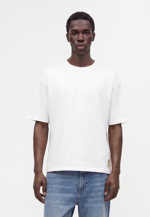 Homme portant un t-shirt blanc uni à manches courtes et un jean bleu, debout avec une expression neutre devant un fond uni.