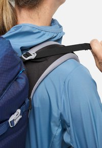 Sac à dos en tissu texturé bleu marine, avec une sangle d'épaule rembourrée noire bordée de mesh gris, porté par une personne vêtue d'une veste bleu clair.