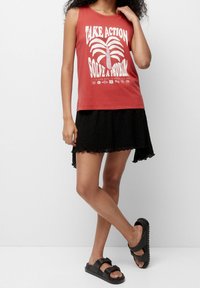 Rød ærmeløs top med hvid tekst og palmegrafik, kombineret med en sort tekstureret nederdel. Sorte sandaler med to remme og tykke såler.