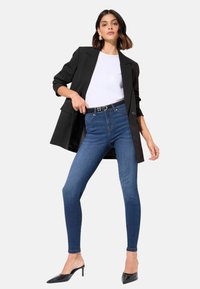 Fekete blézer, fehér póló, kék skinny farmernadrág és fekete hegyes sarkú cipő. A blézernek van egy szerkezeti dizájnja, felgöngyölt ujjakkal. Sima farmer textúra.
