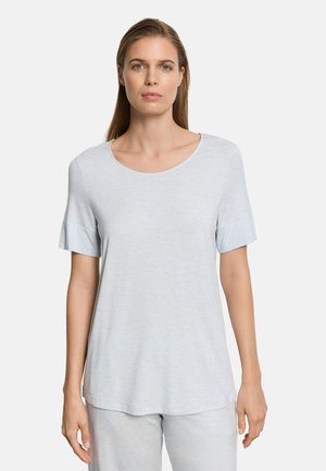 Vrouw met steil haar, gekleed in een lichtblauw T-shirt met korte mouwen en ronde hals, en bijpassende broek, staand tegen een effen achtergrond.