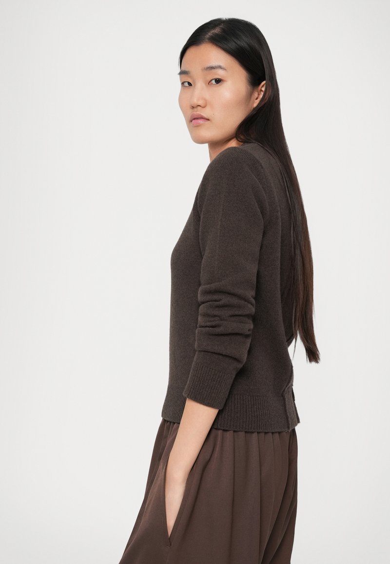Woman with long dark hair wearing a dark brown sweater and matching pants, posing sideways with one hand in pocket against white background.