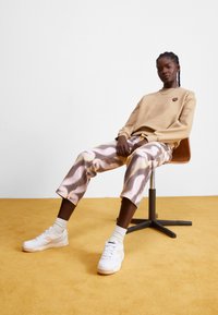Beige sweatshirt with heart logo, patterned pastel pants, and white sneakers. Model seated on a wooden chair, against a plain background.