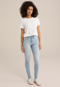 Bela bomba majica s kratkimi rokavi, v kombinaciji s svetlo modrimi skinny jeans hlačami in sivimi športnimi copati z belimi vezalkami. Preprost, vsakdanji outfit.