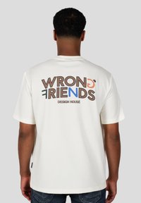 Camiseta de algodón blanca de manga corta, con el texto colorido "AMIGOS EQUIVOCADOS" y "CASA DE DISEÑO" impreso en la parte posterior.