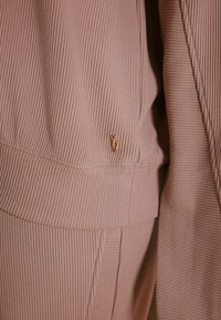 Bežs audums ar ribotu, vertikālu tekstūru, piešūtu mazs zelta akcenta detaļu šuvē. Dizains ir ikdienišķs un pieguļošs.