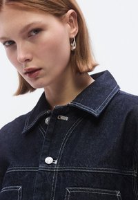 Sötét farmerdzseki világos varrással, ezüstszínű gombokkal és klasszikus gallérral. Simább textúrájú, elülső zsebekkel és testhezálló kialakítással.