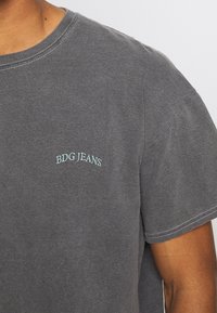 Harmaa puuvillapaita lyhyillä hihoilla, jossa on pyöreä kaula-aukko ja vaaleansinisellä brodeerattu "BDG JEANS" -logo rinnassa.