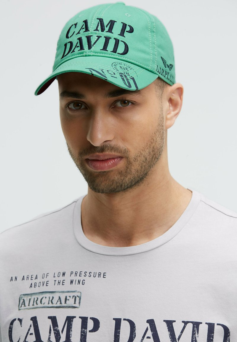 Green baseball cap featuring "CAMP DAVID" in large, dark lettering, with additional graphics. Worn with a light gray T-shirt displaying text.