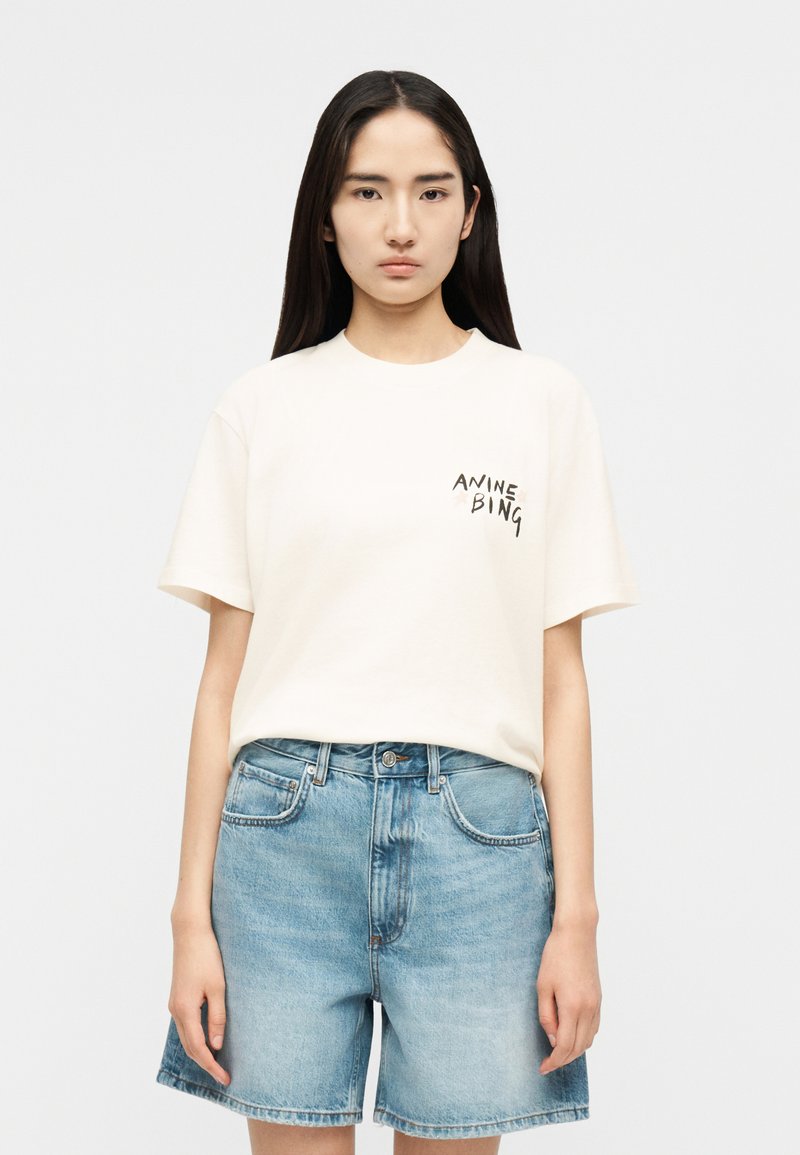 Νεαρή γυναίκα που φοράει λευκό μπλουζάκι με την επιγραφή «ANINE BING» στο στήθος και ανοιχτόχρωμα τζιν σορτς, στέκεται μπροστά σε απλό φόντο.