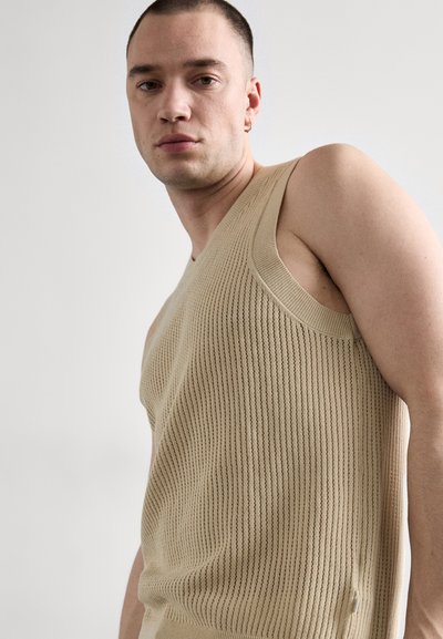 Hombre con cabello corto que lleva un top de punto sin mangas de color beige, mirando directamente a la cámara contra un fondo claro y liso.