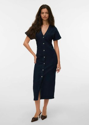 Femme aux longs cheveux bouclés portant une robe midi boutonnée bleu marine à manches courtes et des escarpins marron à bouts pointus, debout devant un fond uni.