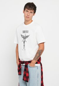 Młody mężczyzna z wytatuowanym przedramieniem, ubrany w białą koszulkę z logo Nirvana "In Utero", niebieskie dżinsy i czerwono-kraciastą koszulę przewiązaną w pasie.