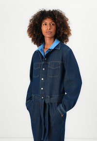 Vrouw met krullend haar, gekleed in een met een riem aangeduide donkere spijkerjas over een blauwe blouse met kraag, staand met haar handen in de zakken, kijkend iets naar rechts.