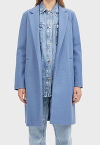 Abrigo de mezcla de lana de color azul claro con un diseño ajustado, solapas en pico y bolsillos laterales, usado sobre una camisa de mezclilla y pantalones vaqueros claros.