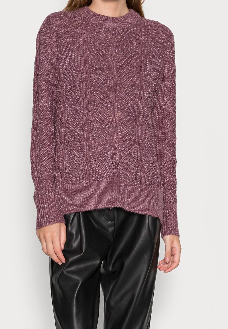Knitted sweater in mauve with intricate cable patterns, ribbed neckline, and cuffs. Paired with loose-fitting black leather pants.