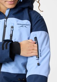 Hellblauer und marineblauer wasserdichter Mantel mit verstellbarem Kapuzen, Reißverschlussarmtasche und Informationsetikett "STX 20.000" an den Ärmeln.