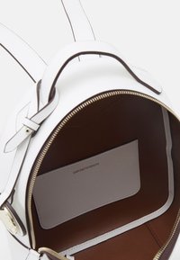 White leather backpack with a textured finish, tan interior, gold zipper, and a small interior pocket displaying the brand name.