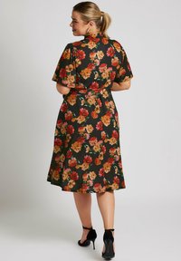 Vestido negro con estampado floral y mangas cortas, acentuado con flores naranjas y rojas, ceñido en la cintura, falda fluida, combinado con tacones altos negros.
