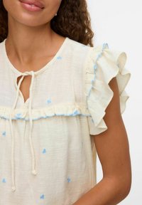 Blusa di un leggero color crema con motivi a cuori blu, maniche a volant, scollatura a chiave con nastri, e un tessuto morbido e strutturato.