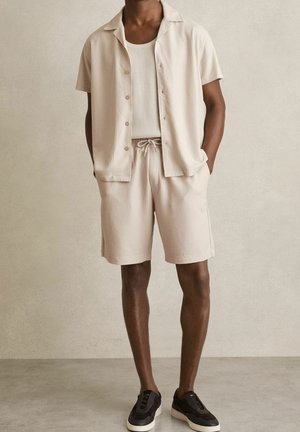 Mann trägt beige Kurzarmhemd, passende Shorts, weißes Tanktop und schwarze Turnschuhe und steht vor einer schlichten beigen Wand.