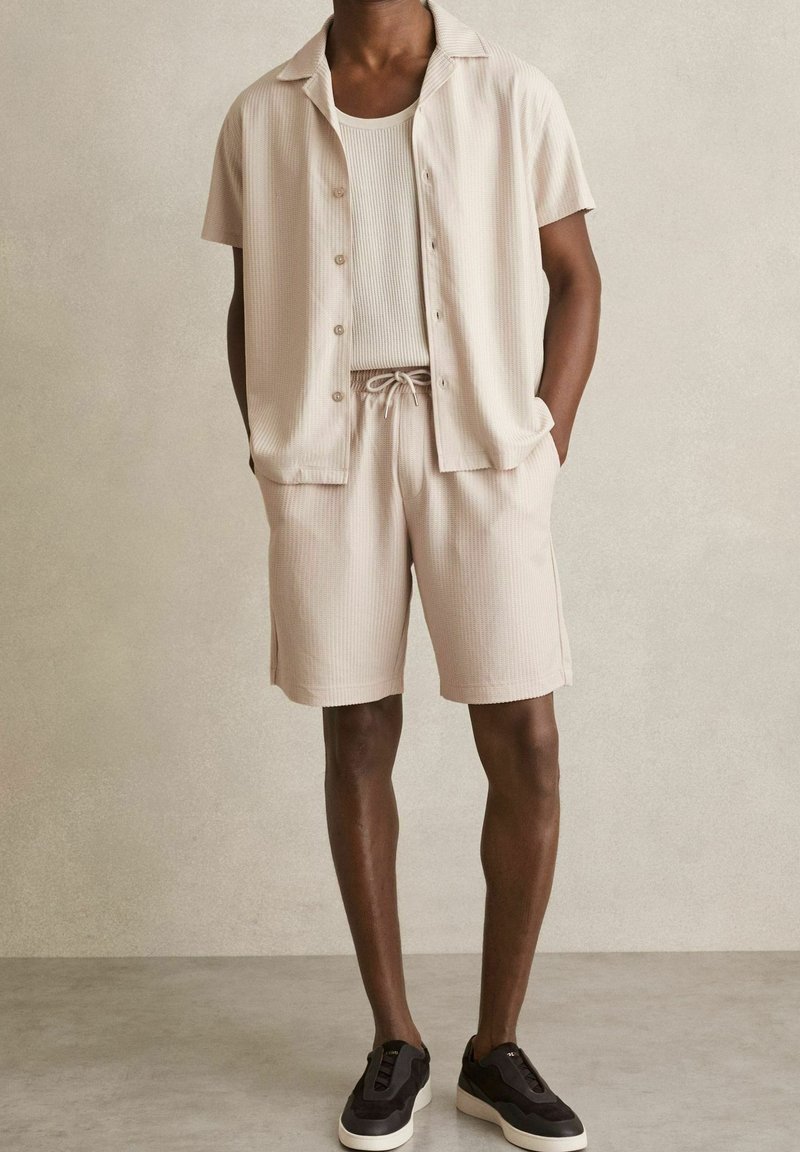 Homme portant une chemise beige à manches courtes, un short assorti, un débardeur blanc et des baskets noires, debout contre un mur beige uni.