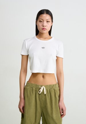Hvid cropped t-shirt lavet af ribbet materiale med korte ærmer og et lille sort logo på brystet, parret med grønne kordfløjl bukser.
