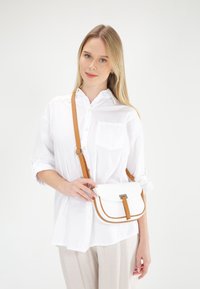 Biała torebka crossbody z teksturowanym wykończeniem, brązowymi akcentami, prostokątnym kształtem i srebrnymi dodatkami. Noszona z koszulą z guzikami.