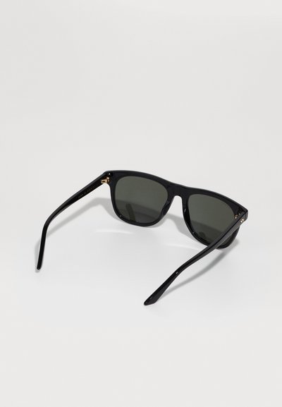 Ochelari de soare din plastic negru cu lentile întunecate, formă pătrată și brațe subțiri. Prezentând accente de hardware aurie lângă balamale.