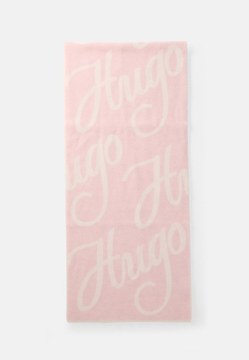 Lyserød tæppe med stort hvidt "Hugo"-tekst, blød tekstur, rektangulær form og subtile frynsekant. Lavet af hyggeligt materiale.