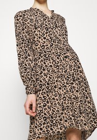 Leoparddruckkleid in Beige mit schwarzen Flecken, mit V-Ausschnitt, langen Ärmeln und einer fließenden Silhouette, die in der Taille geschnitten ist.