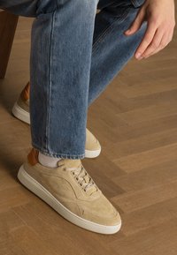 Beige suede sneakers with a textured finish, featuring a tan leather accent on the heel, cream rubber sole, and flat laces. Worn with blue jeans.