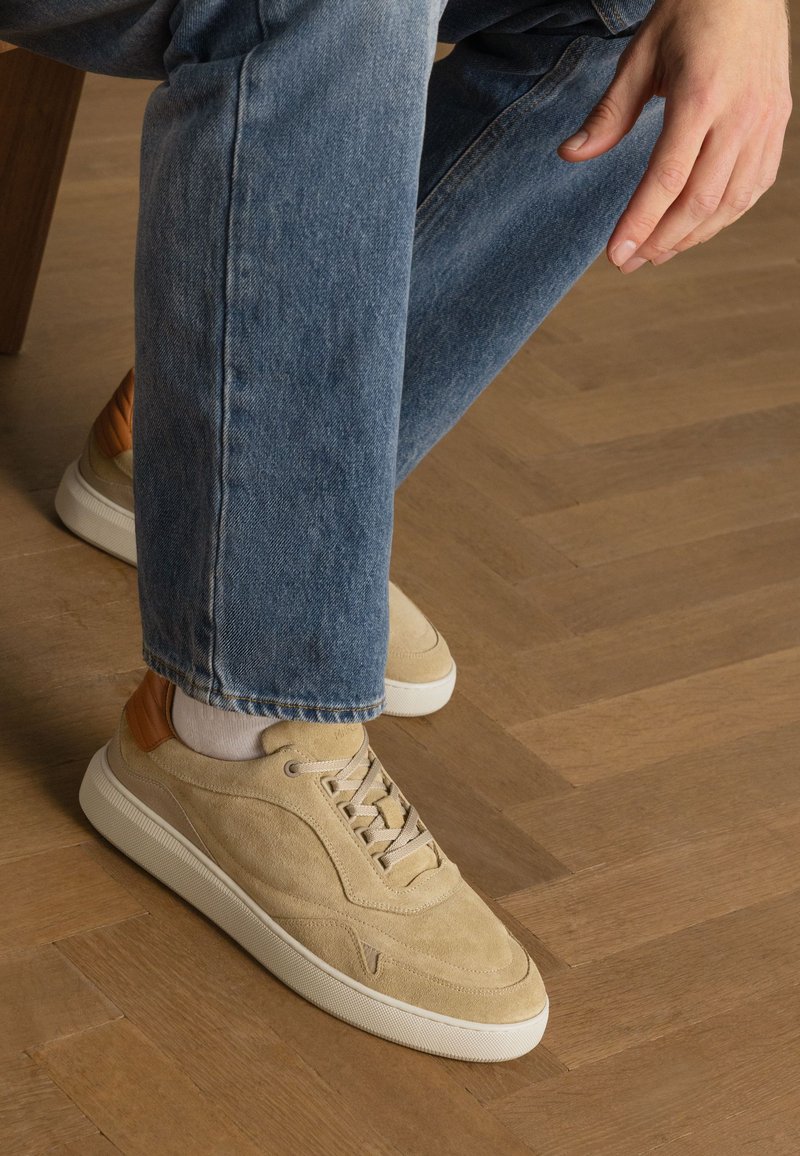 Beige suede sneakers with a textured finish, featuring a tan leather accent on the heel, cream rubber sole, and flat laces. Worn with blue jeans.
