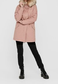 Parka rose clair avec capuche en fausse fourrure, fermeture éclair devant et deux poches latérales. Associée à un pantalon noir et des bottes noires à lacets.