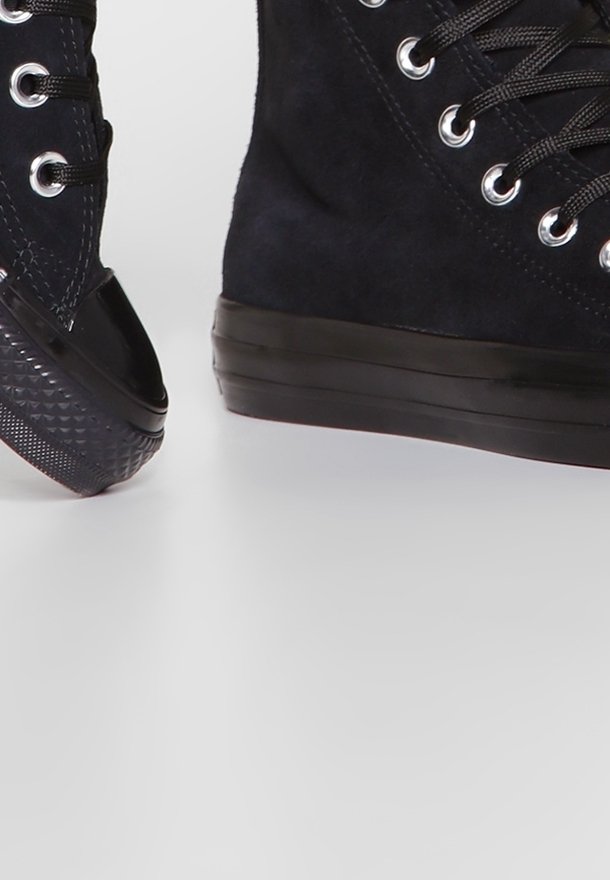 Czarne sneakersy za kostkę z zamszowymi cholewkami, czarnymi gumowymi noskami, czarnymi sznurowadłami i grubymi czarnymi gumowymi podeszwami na jednolitym tle.