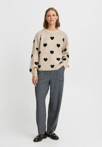 Beige Strickpullover mit schwarzen Herzmustern, mit langen Ärmeln. Graue maßgeschneiderte Hosen mit Fronttaschen, kombiniert mit schwarzen Loafers.