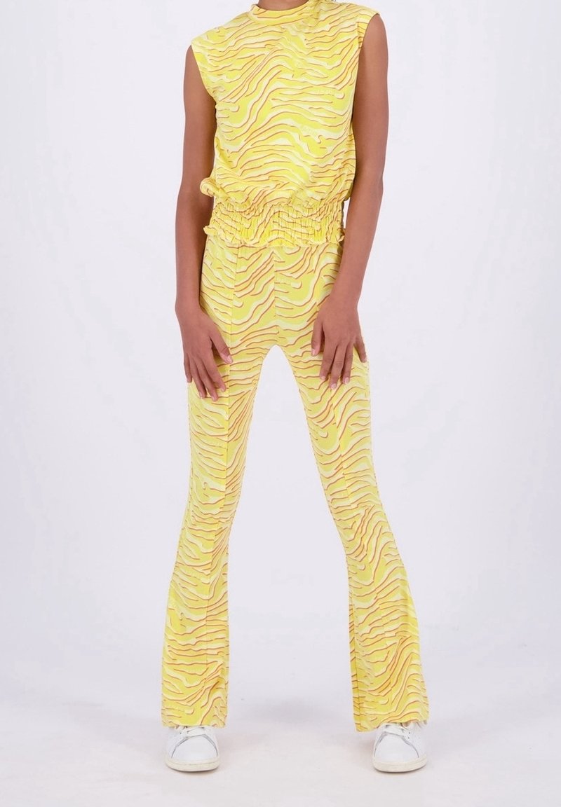 Personne portant un haut jaune sans manches et un pantalon évasé assorti avec des rayures brunes ondulées, debout les mains détendues le long du corps.