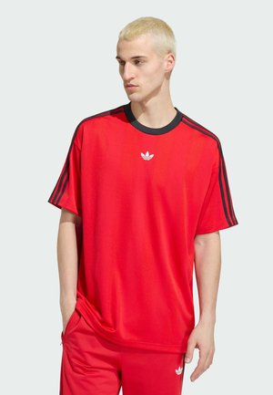 Blond muž v červeném tričku Adidas s černými pruhy na rukávech a odpovídajících červených kalhotách s logy Adidas, stojící před jednobarevným pozadím.
