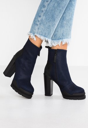 Femme portant des bottines bleu foncé à talons carrés et fermeture éclair latérale, associées à un jean bleu clair à ourlet effiloché.