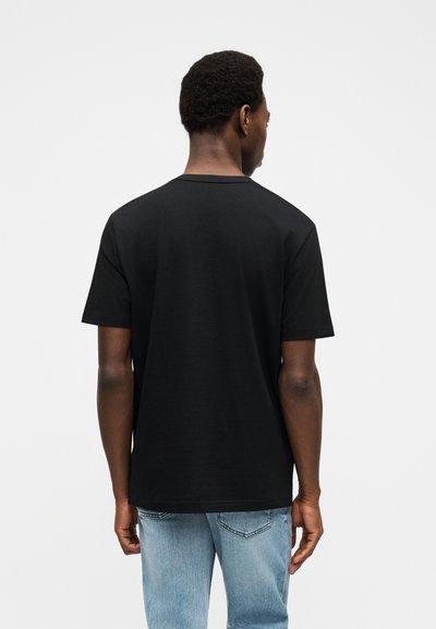 Homme aux cheveux courts et bouclés portant un t-shirt noir uni à manches courtes et un jean bleu clair, vu de dos sur fond blanc.