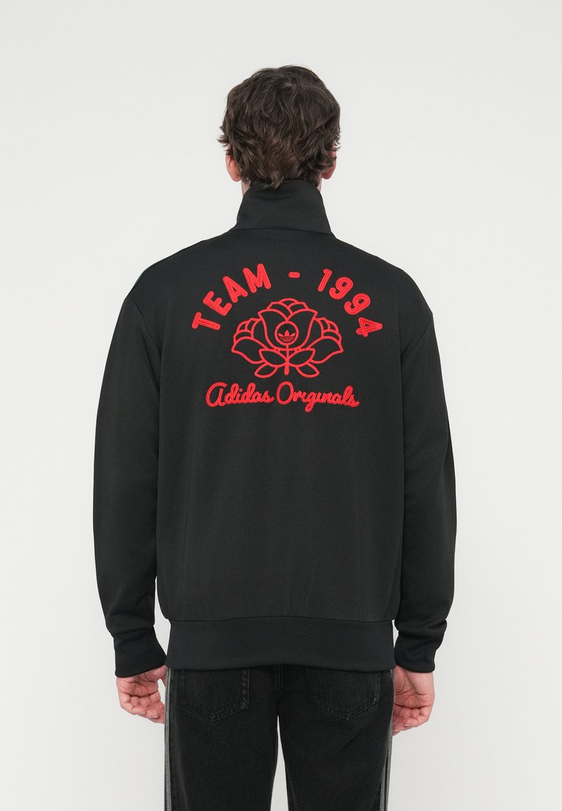 Sort sweatshirt med høj krave, rød broderet tekst "TEAM - 1994" og et rosedesign med "Adidas Originals." Ensartet tekstur.