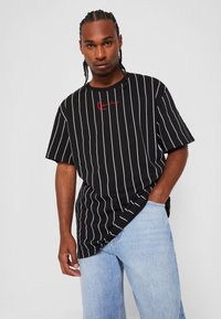 T-shirt noir à rayures avec des lignes verticales blanches, arborant un logo rouge. Le tissu est doux, avec une coupe décontractée et un design à épaule tombante.