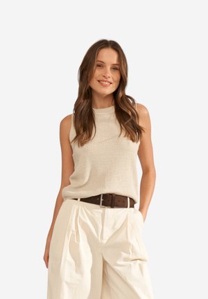 Femme souriante aux longs cheveux bruns portant un haut beige sans manches en tricot, rentré dans un pantalon crème taille haute avec une ceinture foncée.