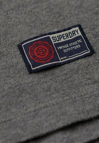 Tela gris con una textura tejida, que presenta un parche azul y rojo que dice "Superdry Vintage Athletic Outfitters". Costura en los bordes visible.