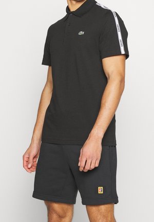 Mężczyzna noszący czarną koszulkę polo Lacoste z białą taśmą z logo na rękawach oraz czarne szorty z małą prostokątną naszywką na lewej nodze.