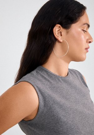 Boucle d'oreille en argent avec une surface lisse, suspendue à l'oreille d'une femme. Haut sans manches gris avec une texture côtelée. Fond neutre.