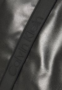 Tecido preto texturizado com um acabamento brilhante. Apresenta uma alça preta proeminente com a inscrição "Calvin Klein" ao longo de seu comprimento.