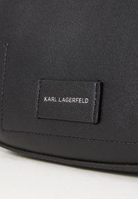 Mustakotelo, jossa on sileä pinta, pyöreä muoto ja näkyvä etiketti, johon on kohokuvioitu "KARL LAGERFELD" valkoisella.