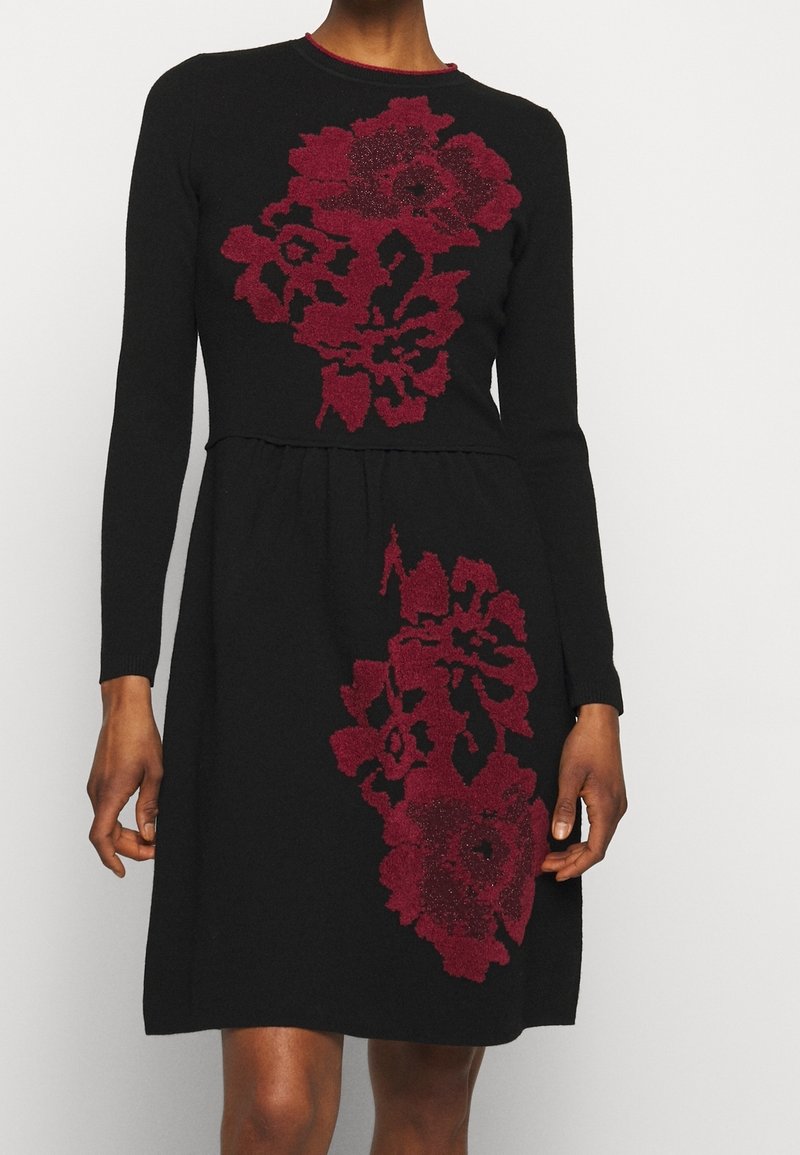 Robe noire à manches longues avec motifs floraux rouges sur la poitrine et la jupe, portée par une personne debout sur un fond uni.
