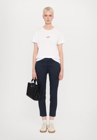 Hvid bomuld t-shirt med sort logo, marineblå slim-fit bukser, beige sneakers og flettet sort håndtaske med korte håndtag. Ensfarvet baggrund.